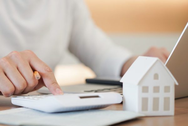 Questions to Help Set Home Replacement Cost - Homeowner Calculating Potential Risk Costs Using Laptop and Calculator with A White Desk Ornament in the Shape of a Home on Their Desk