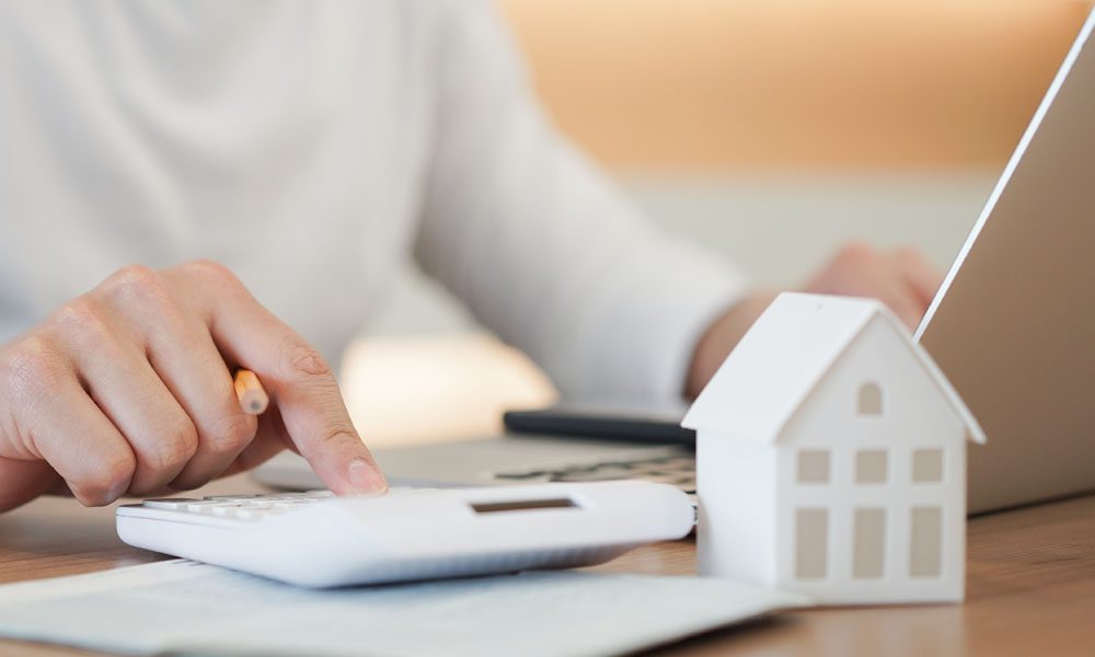 Questions to Help Set Home Replacement Cost - Homeowner Calculating Potential Risk Costs Using Laptop and Calculator with A White Desk Ornament in the Shape of a Home on Their Desk